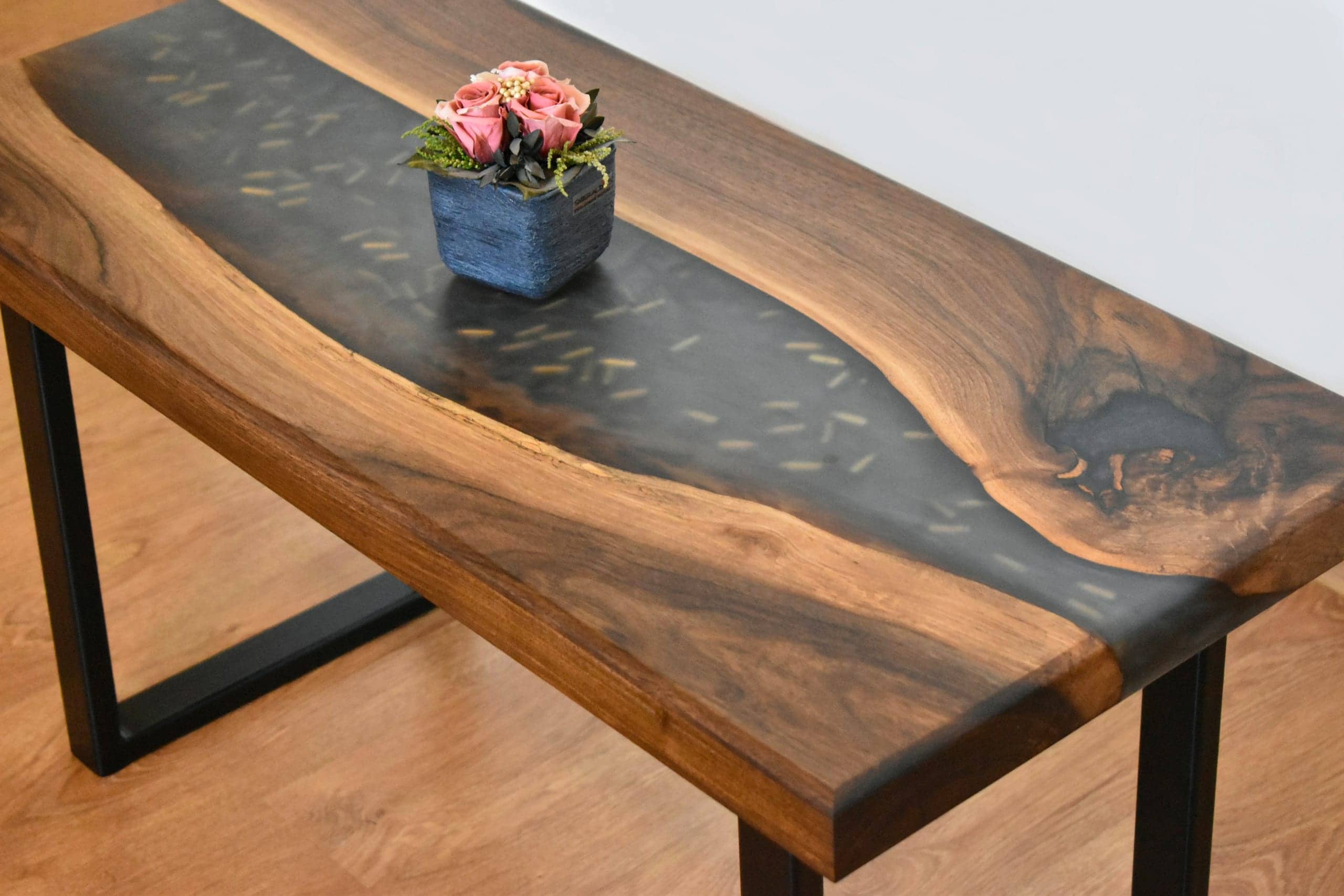 Ein Holztisch mit einem dunklen Fluss aus Epoxidharz mit goldenen Einschlüssen, darauf ein blauer Topf mit Rosen.