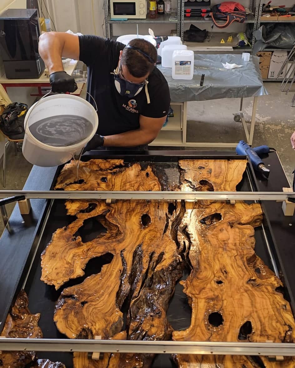 Michael Scabell giesst mit Atemschutzmaske Epoxidharz aus einem Eimer über Olivenholzplatten in einer Giessform.