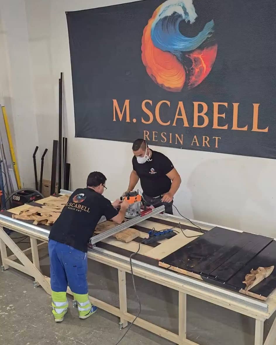 Zwei Männer fräsen Holzplatten auf einem langen Werktisch, dahinter hängt ein grosses Banner mit dem Logo von M. Scabell Resin Art.